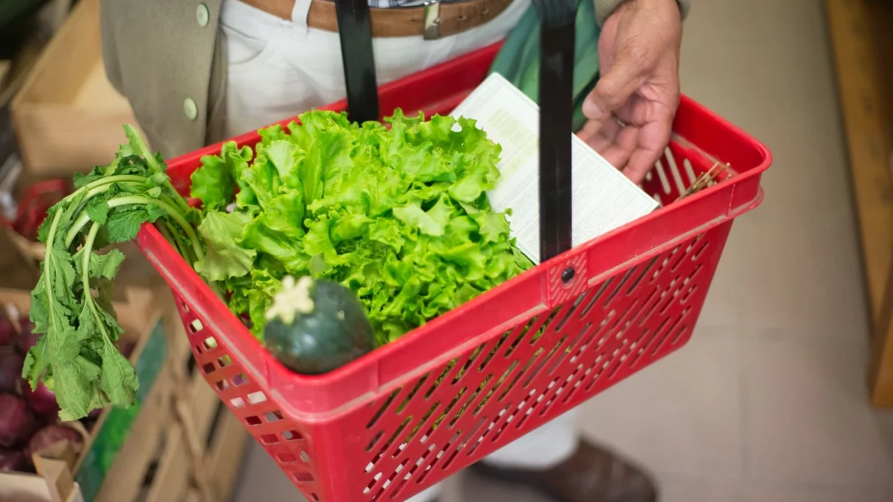 A hand carrying a red grocery basket filled with vegetables